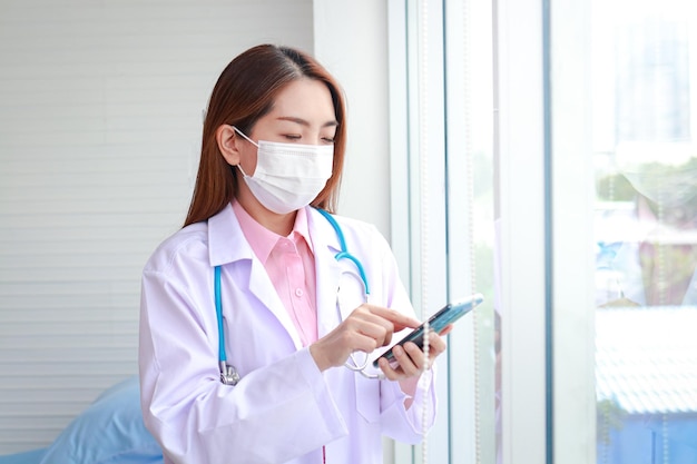 Beautiful Asian female doctor wearing a white mask stand holding a smartphone chat online through the application Modern communication technology concept Patient care through smartphones