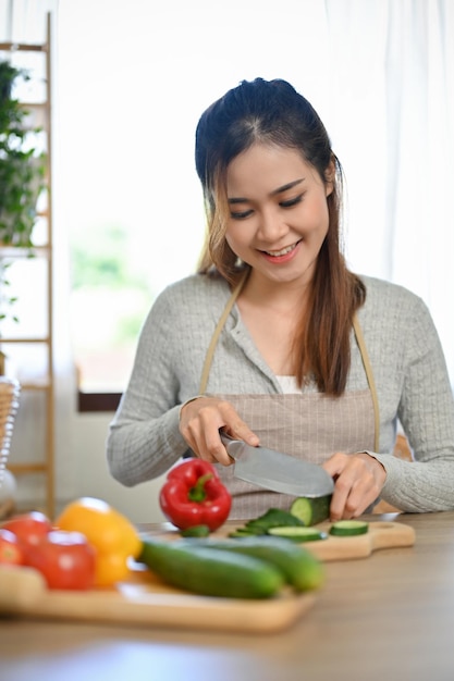 Beautiful Asian female housewife wearing apron chopping cucumber in the kitchen