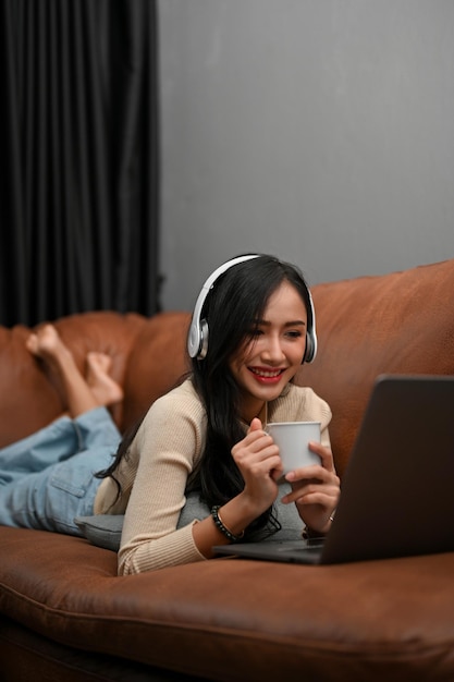 Beautiful Asian female lying on sofa watching movie on her laptop and sipping coffee