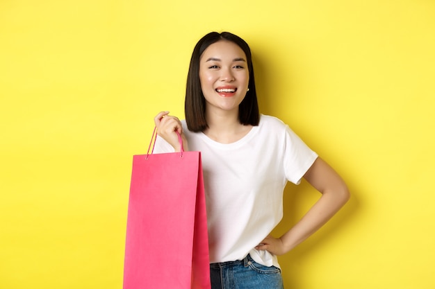 Beautiful asian female model in white t-shirt, holding shopping bag and smiling satisfied, standing over yellow.