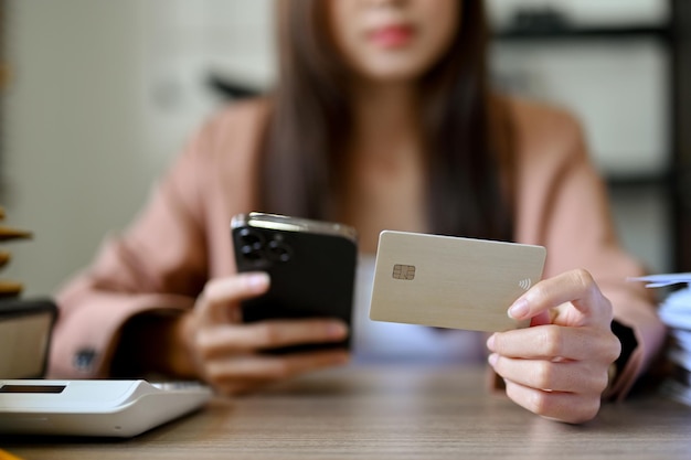 A beautiful Asian female sits at her desk holding a credit card and a smartphone