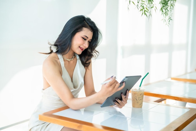 Beautiful asian freelance Success people business woman fashion model in summer white sleeveless dress using casual working with tablet for browsing internet chatting and blogging in coffee shop