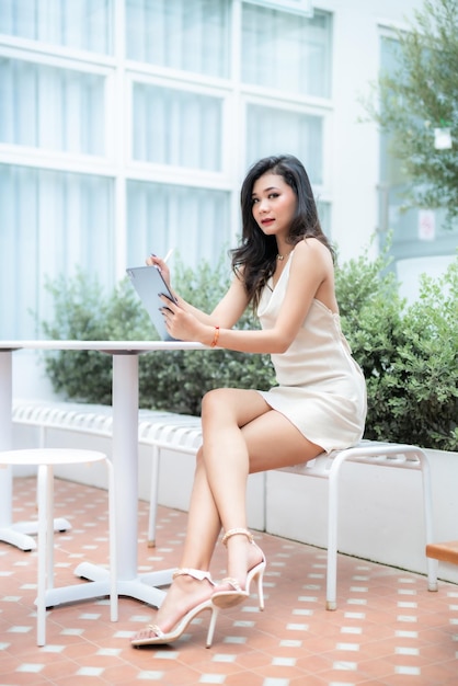 Beautiful asian freelance Success people business woman fashion model in summer white sleeveless dress using casual working with tablet for browsing internet chatting and blogging in coffee shop