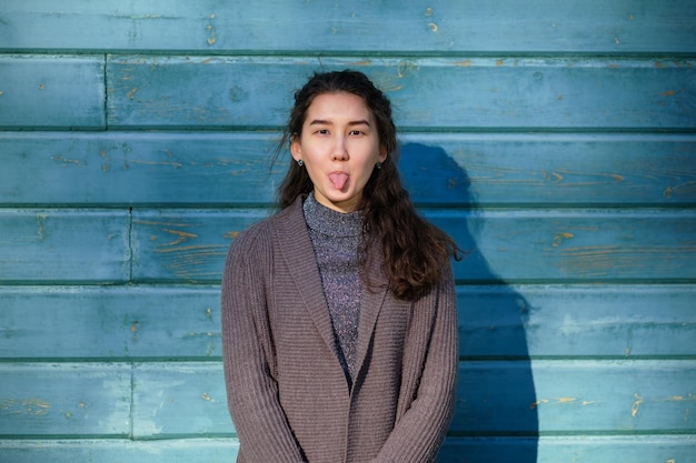 A beautiful Asian girl fools around and shows her tongue She stands by the wooden blue wall She wears a warm jacket