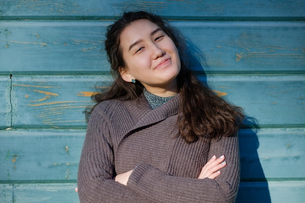 A beautiful Asian girl with long hair and a warm sweater is standing by the wooden blue wall She looks forward with smile