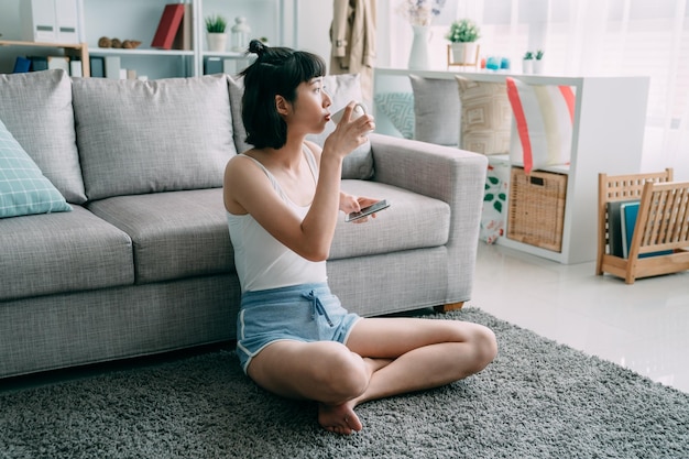 Beautiful asian korean girl in home clothes using smartphone while sitting on carpet in home living room. young charming woman enjoy leisure time in weekend morning. lady hold mobile phone drink tea