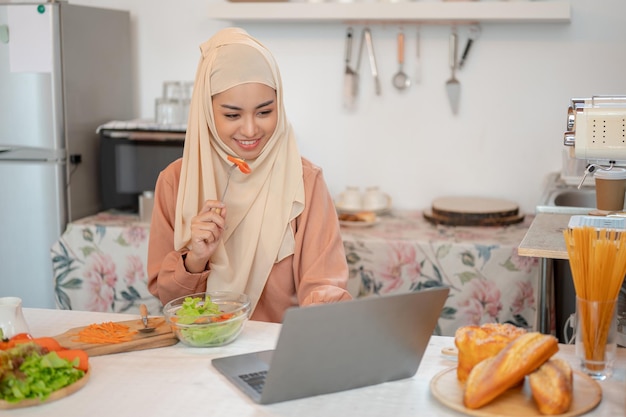 A beautiful Asian Muslim woman eating her healthy yummy salad bowl while using her laptop