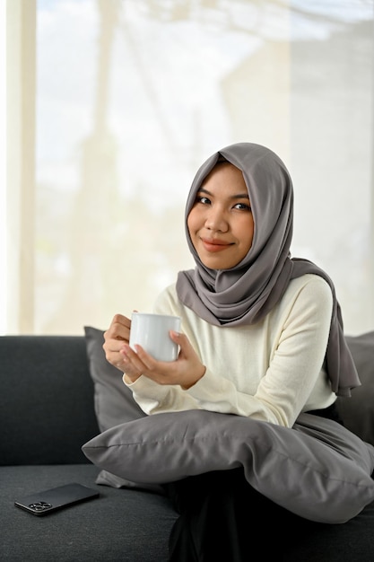 Beautiful Asian Muslim woman in hijab holding a coffee cup sitting on sofa in the living room