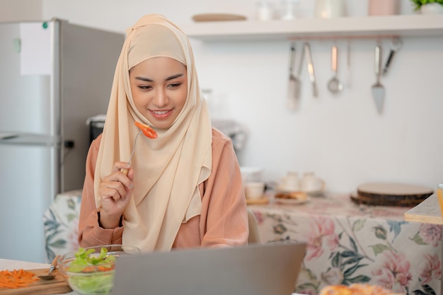 A beautiful Asian Muslim woman is eating her salad bowl while working on her laptop