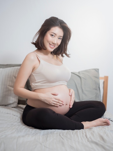 Beautiful asian pregnant woman sit on bed and touching her belly 