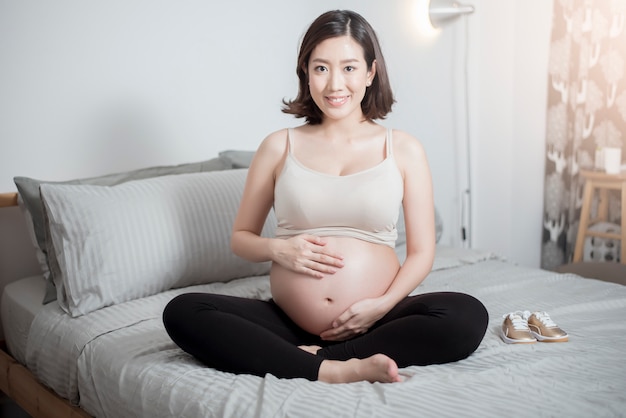 Beautiful asian pregnant woman sit on bed and touching her belly 