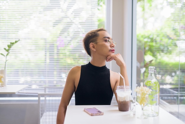 Beautiful Asian queer man sitting in coffee shop and looking through window while thinking