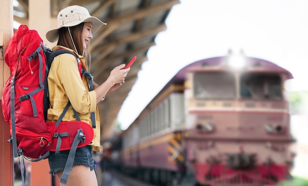 Beautiful asian traveler with backpack using smartphone while standing at railway station and waiting for departure