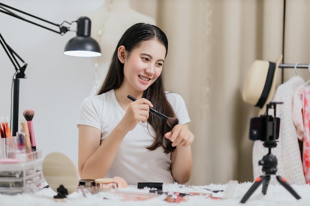 Beautiful Asian woman blogger showing how to make up and use cosmetics live streaming online to social media at home with a smartphone Beauty blogger or influencer online concept