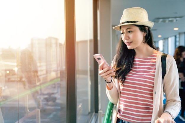 beautiful asian woman browsing her smartphone standing next to large windows.