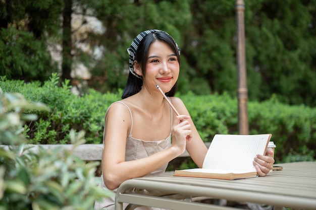 A beautiful Asian woman in a cute dress is daydreaming while writing her diary in the green garden