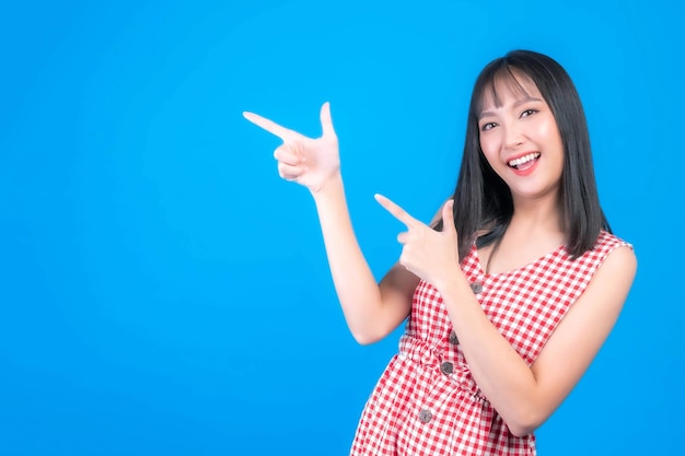 Beautiful Asian woman cute girl with bangs hair style in blue t shirt smiling and pointing finger to empty copy space for present product or blank space for advertising isolated on blue background