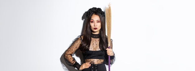 Beautiful asian woman in gothic lace dress and black wreath holding broom and looking suspicious at