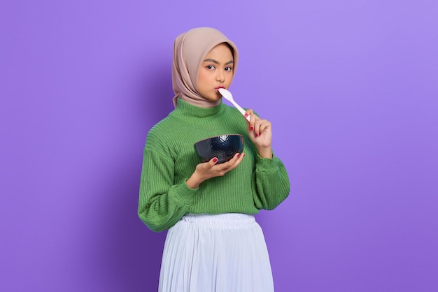 Beautiful Asian woman in green sweater and hijab bowl with cereal and licking a spoon isolated over purple background