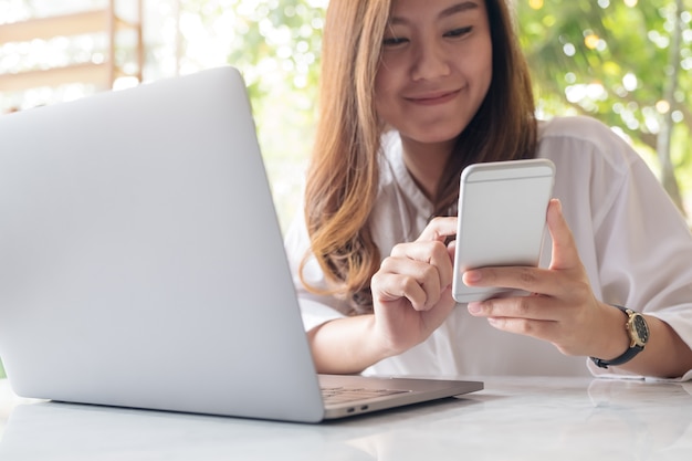 a beautiful Asian woman holding and looking at smart phone while using laptop in cafe