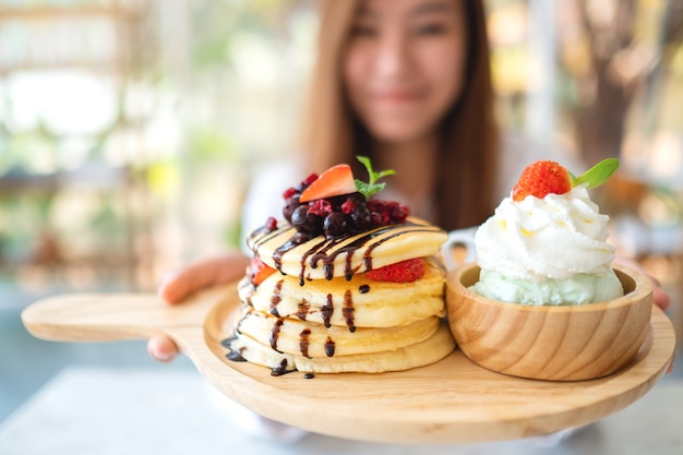 A beautiful asian woman holding and showing a plate of pancakes with ice cream and whipped cream in cafe