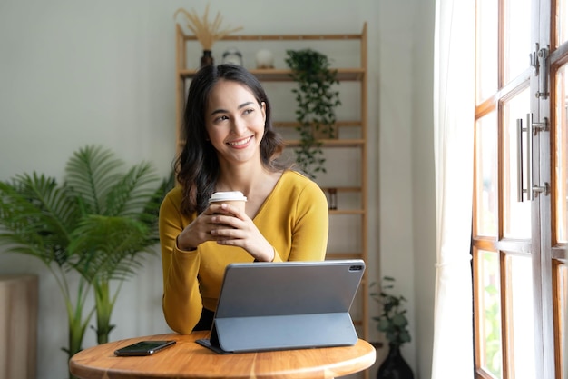 Beautiful Asian woman is using laptop drinking coffee and smiling while sitting by the window at home