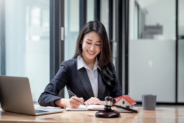 Beautiful Asian woman lawyer taking notes work on real estate loan contract example  home of a mallet with a laptop placed at the office.