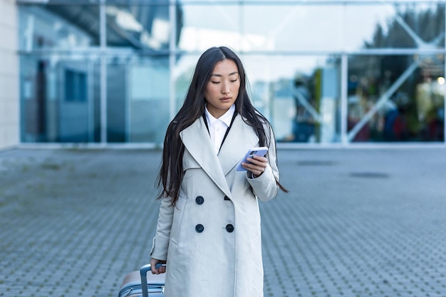 Beautiful Asian woman, near the airport uses a phone application for online booking, a tourist with a large suitcase