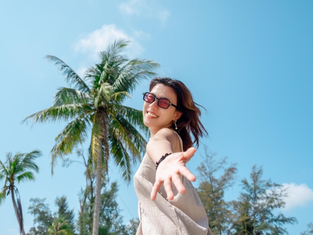 Beautiful Asian woman portrait with sunglasses on the beach in summer vibes Happy female smile looking at camera in front of tropical palm trees on blue sky background Holiday vacation summertime