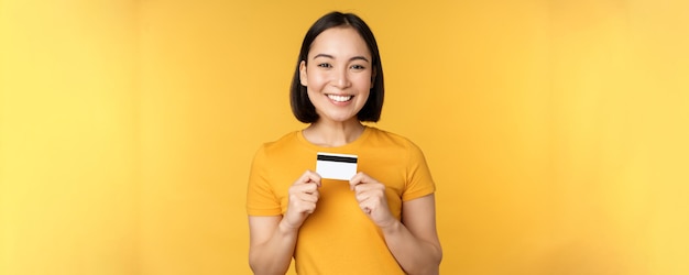 Beautiful asian woman showing credit card and smiling recommending bank service standing over yellow background Copy space