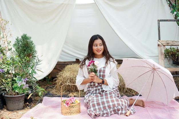 Beautiful Asian woman sitting on picnic mat with artificial flower in vintage style countryside farm