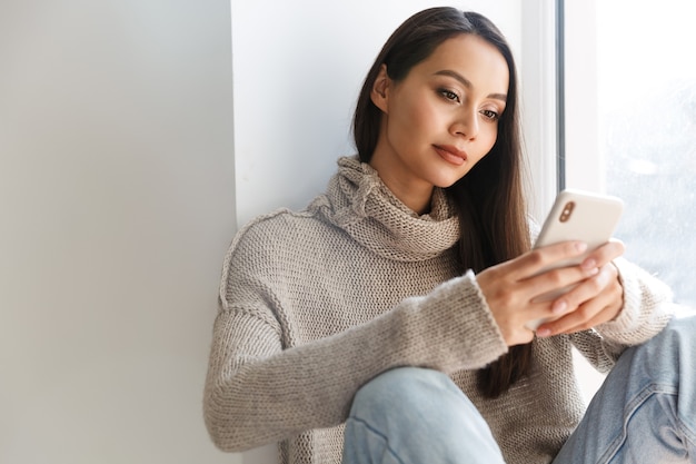 beautiful asian woman typing on mobile phone while sitting on window sill indoors