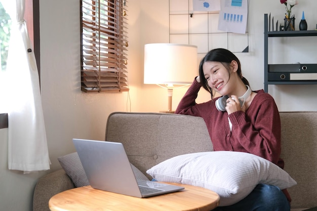 Beautiful Asian woman using laptop while headphones sitting on sofa at home