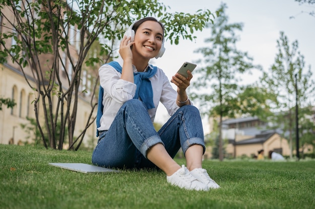 Beautiful asian woman using mobile phone, listening to music outdoors, sitting on grass in park