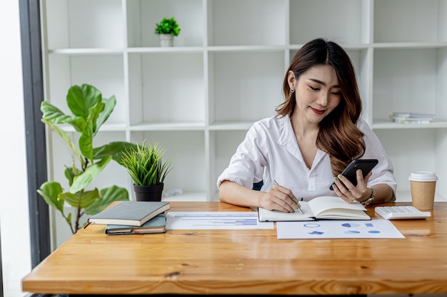 Beautiful Asian woman using a smartphone, businesswoman searching internet on smartphone and taking notes in a notebook, she is working in her room. Concept of Internet search and use of technology.