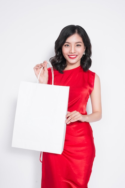 Beautiful asian woman wearing a red dress with shopping bag standing over white.