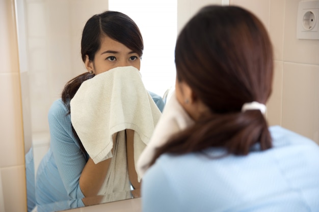 Beautiful asian woman wiping her face with towel, looking at mirror