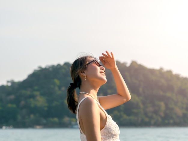 Beautiful Asian woman with sunglasses on the beach in summer vibes Happy smile female covering the sunshine with her hand on the sea green mountain island and sky background Holiday vacation