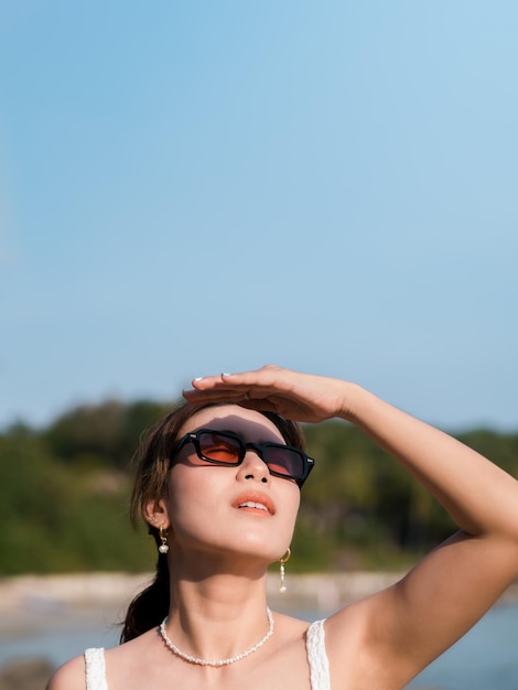 Beautiful Asian woman with sunglasses on the beach in summer vibes with copy space Attractive female covering the sunlight with hand at the sea and sky background Holiday vacation vertical style