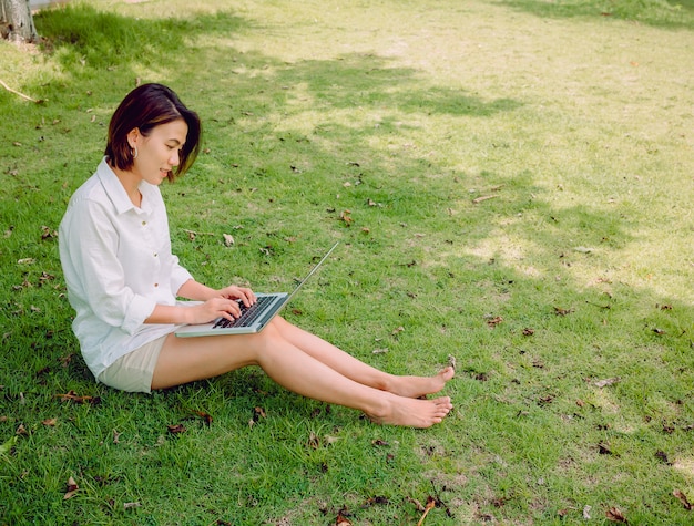 Beautiful Asian women wearing white shirt and shorts with laptop computer sitting on green grass.