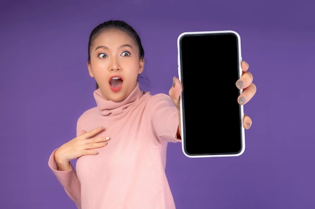 Beautiful Asian young woman Excited surprised girl showing smart phone with blank screen white screen isolated on purple background Mock Up Image