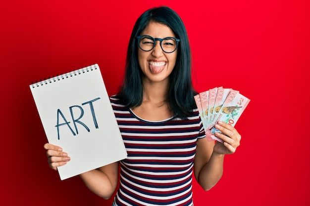 Beautiful asian young woman holding art notebook and 100 new zealand dollars sticking tongue out happy with funny expression