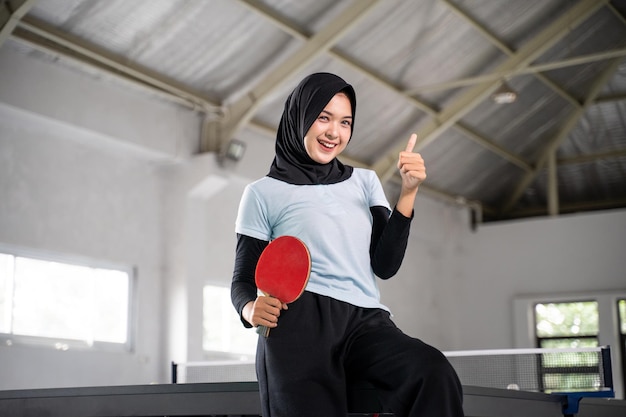 Beautiful athlete in hijab holding paddle with thumbs up inside building