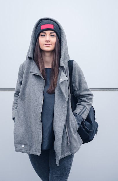 Beautiful athletic girl with dark hair with bennie, a hoodie and a backpack on white wall background