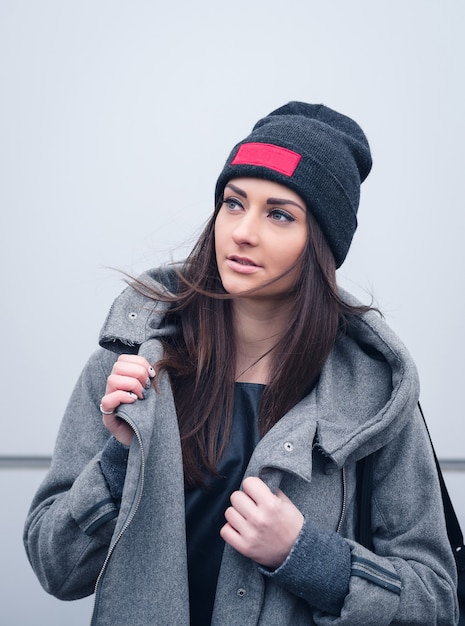 Beautiful athletic girl with dark hair with bennie, a hoodie and a backpack on white wall background