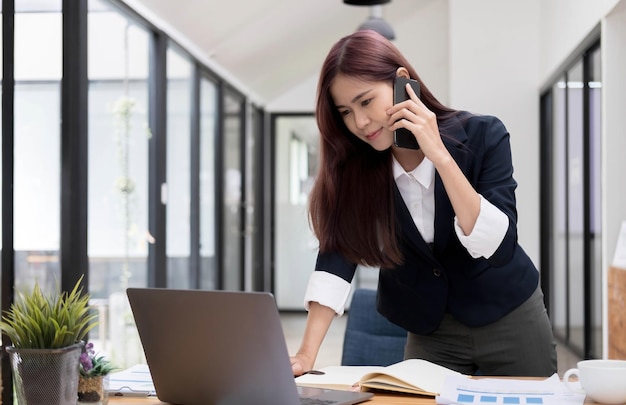 Beautiful attractive young asian businesswoman talking on the phone with her colleague and checking the informations on laptop screen