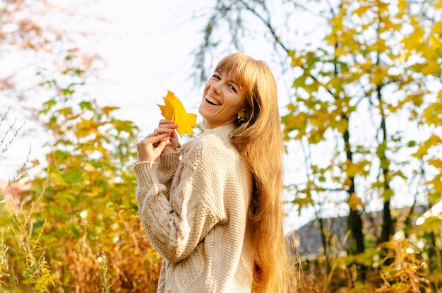 Beautiful autumn woman standing near colorful autumn leaves Pretty happy cheerful model looking at camera Young woman with autumn leaf in hand autumn mood relaxation walking in nature