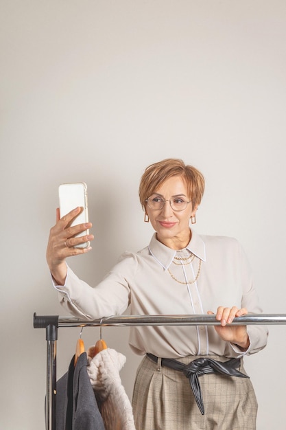 Beautiful baby boomer woman of middle age in glasses smile and makes selfie on smartphone fashion st...