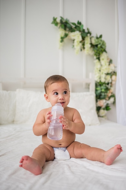 A beautiful baby boy in a diaper sits on the bed and looks away with a bottle of water