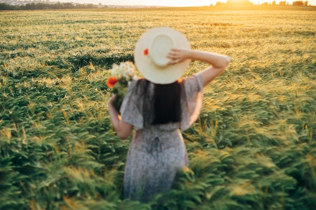 Beautiful barley field in sunset light and blurred image of woman in straw hat holding wildflowers Evening summer countryside and gathering flowers Atmospheric tranquil moment